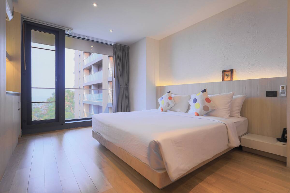 Un dormitorio con una cama grande, un sofá, una ventana con vistas a edificios y una lámpara en el techo.