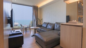 A modern living room with a sofa, a TV, a dining table, and a view of the sea.