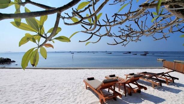 A beach with white sand, wooden lounge chairs, and a view of the sea with boats.