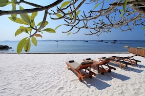 En strand med hvidt sand, træloungestole og udsigt til havet med både.