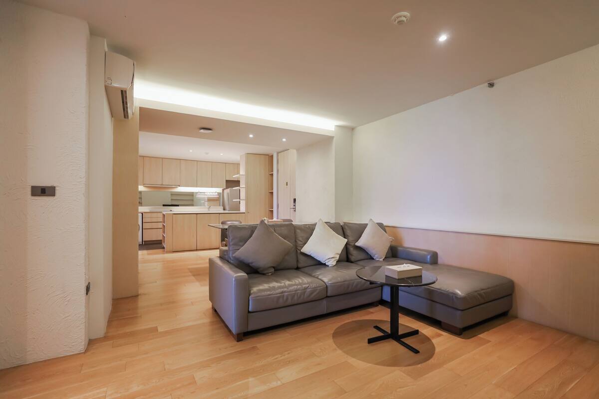 Una sala moderna con un sofá seccional gris, una mesita redonda y una cocina al fondo.