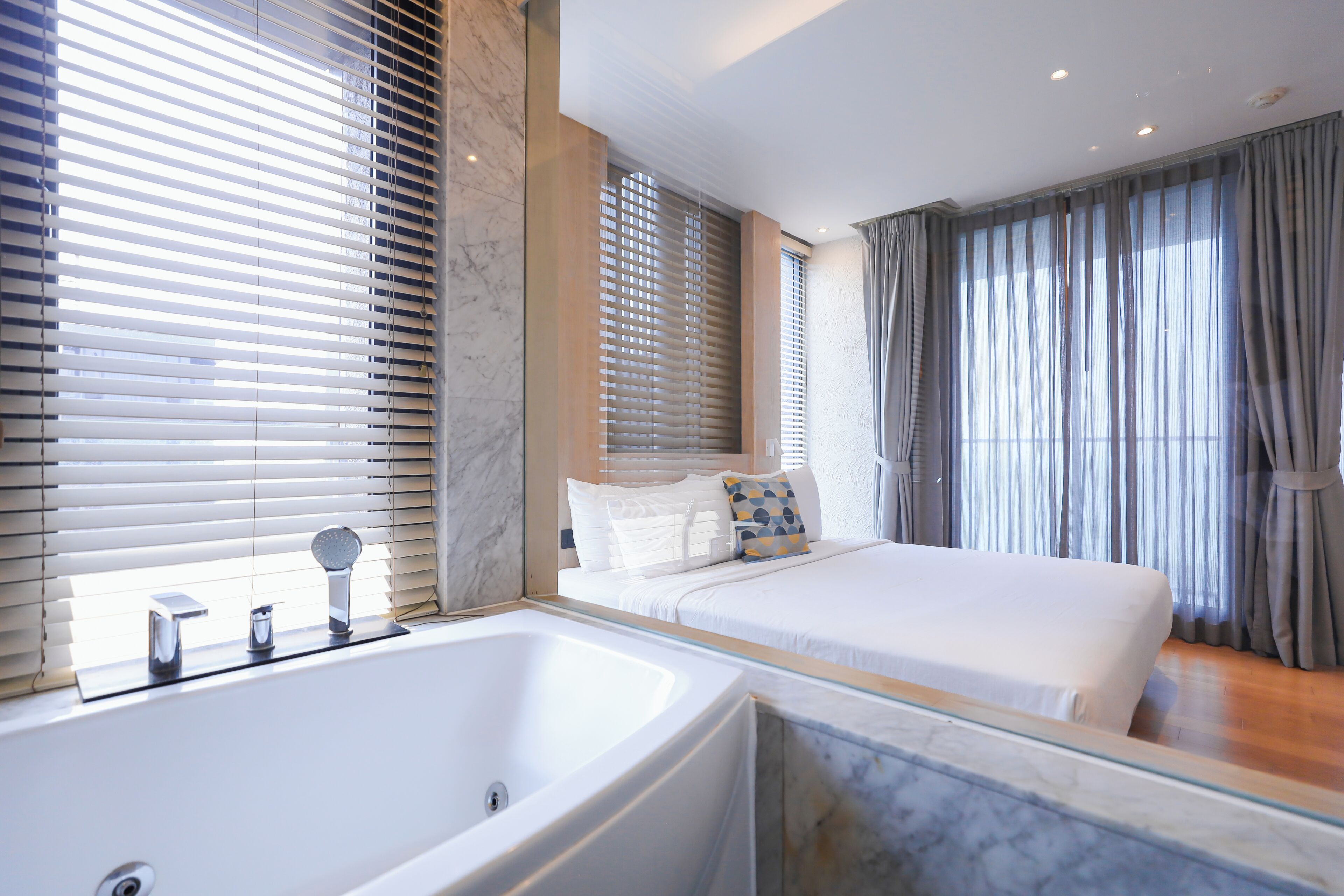 A modern hotel room with a large bathtub, a bed with white bedding, and a large window with blinds.
