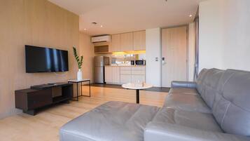 A modern living room with a sofa, a flat-screen TV mounted on the wall, a small table, and a kitchen area in the background.