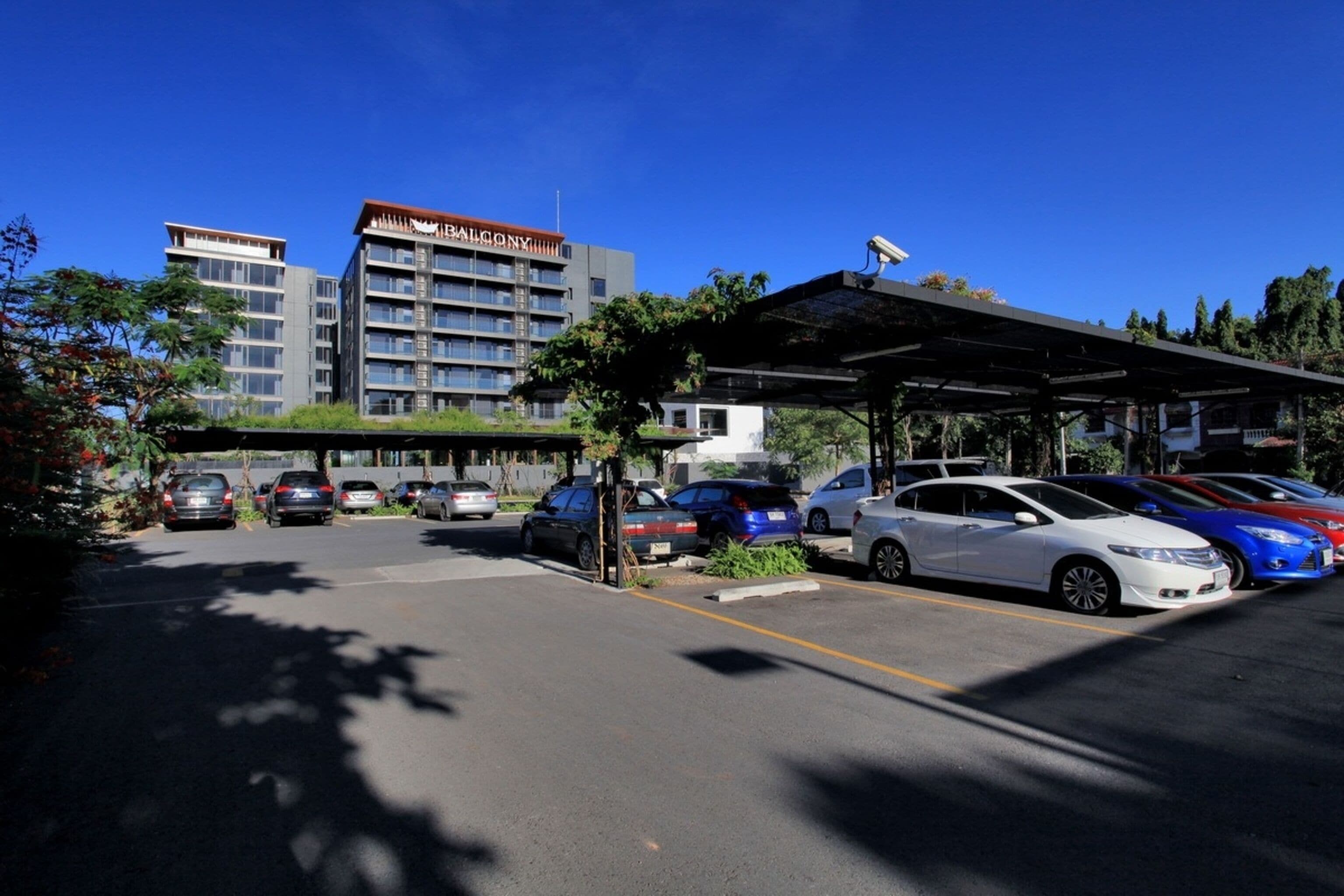 Un estacionamiento con varios autos, una zona de espera techada y un edificio con balcones al fondo.