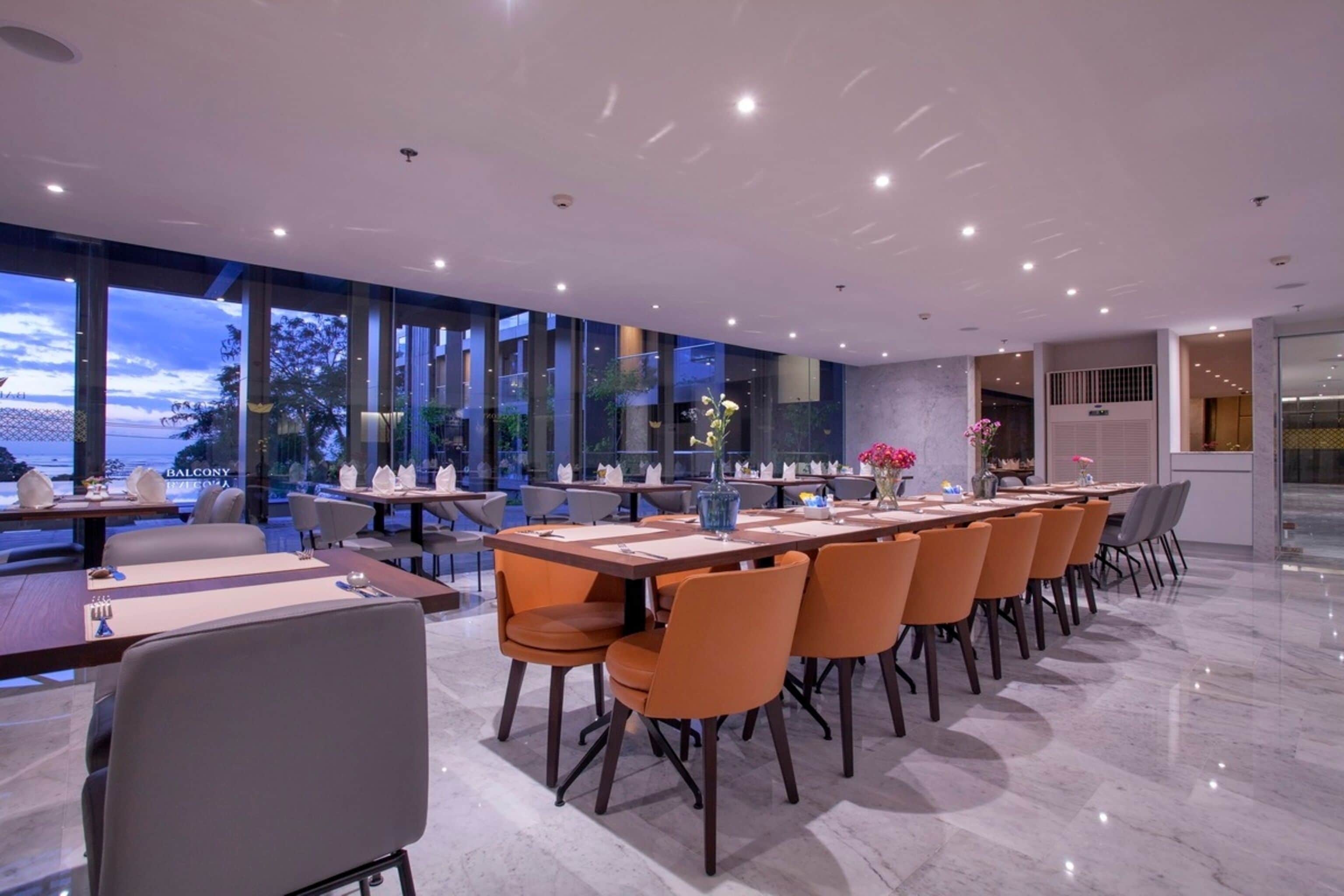 A modern dining area with long tables and orange chairs, large windows offering a view of the outdoors, and a clean, contemporary design.