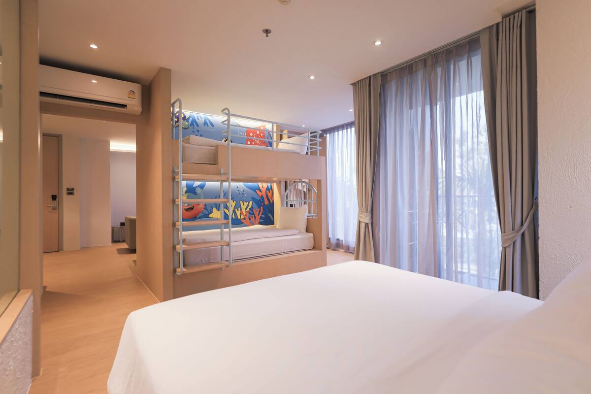 Habitación de hotel con literas, una cama, una ventana con cortinas, un equipo de aire acondicionado y una puerta que da a otra habitación.