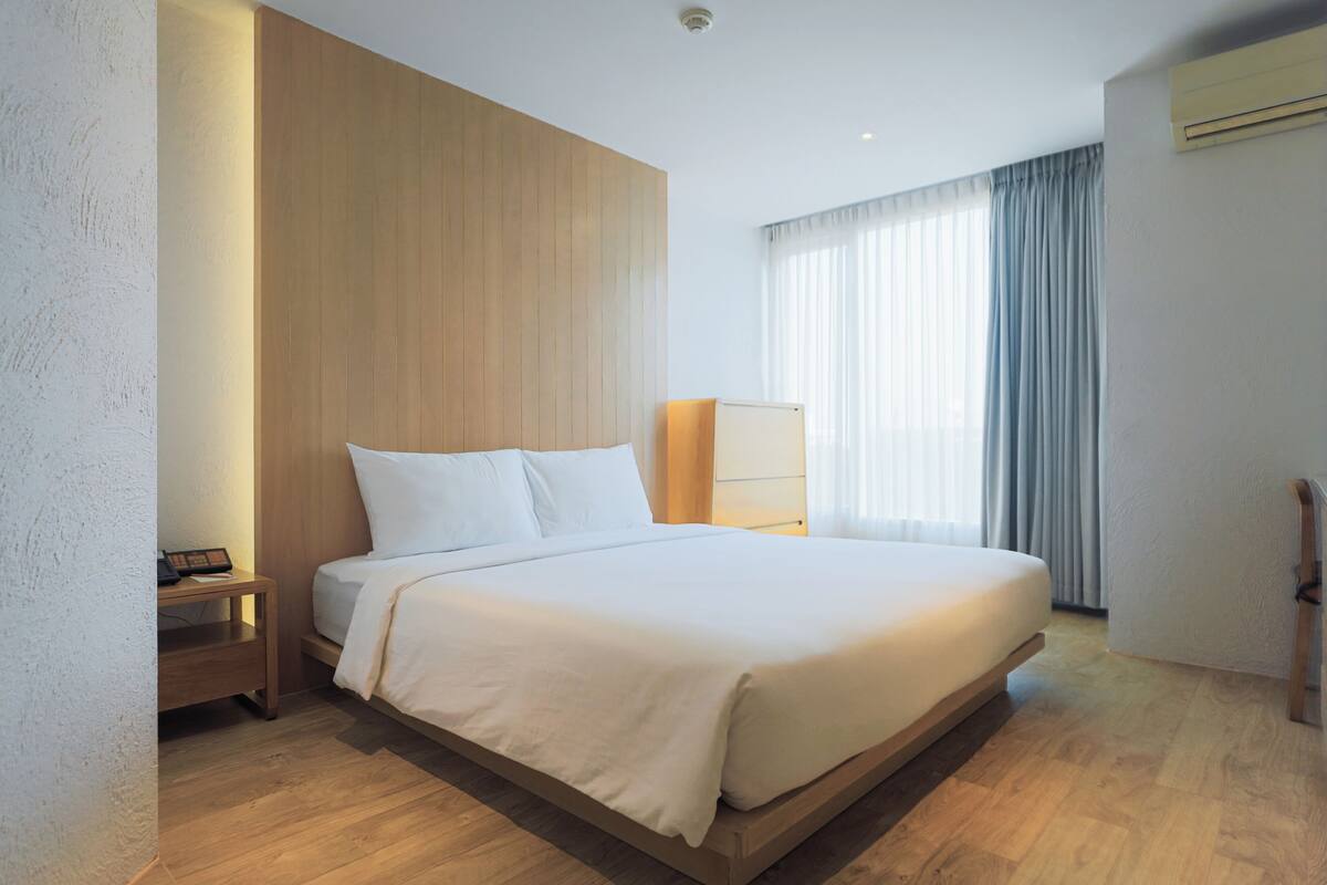Una habitación de hotel moderna con una cama grande, una mesita de noche, una lámpara y una ventana con cortinas.