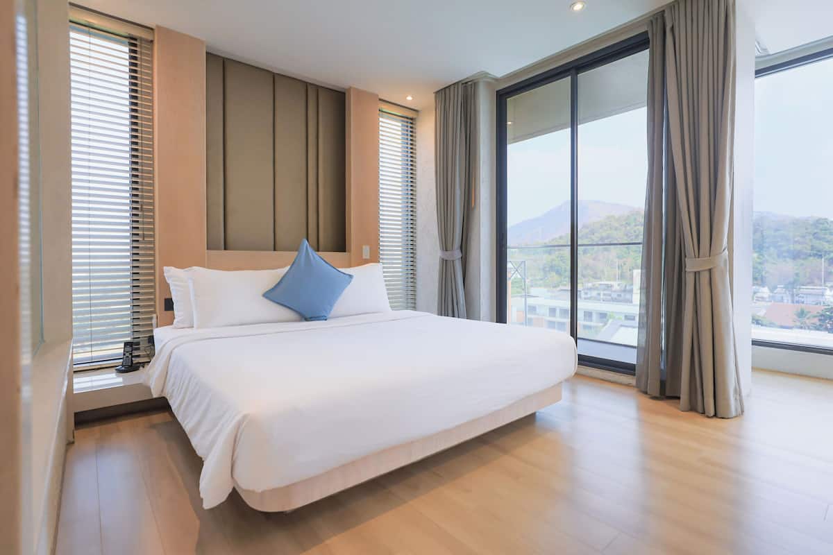 Una cama grande con ropa de cama blanca y un cojín azul, una ventana amplia con cortinas y vistas a un paisaje urbano.