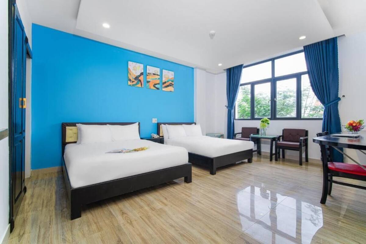 A hotel room with two beds, a dining table, chairs, a small table with a vase of flowers, and artwork on the wall.
