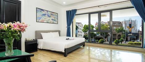 A modern hotel room with a large bed, a desk with a vase of flowers, a painting on the wall, and a view of the city through large windows.