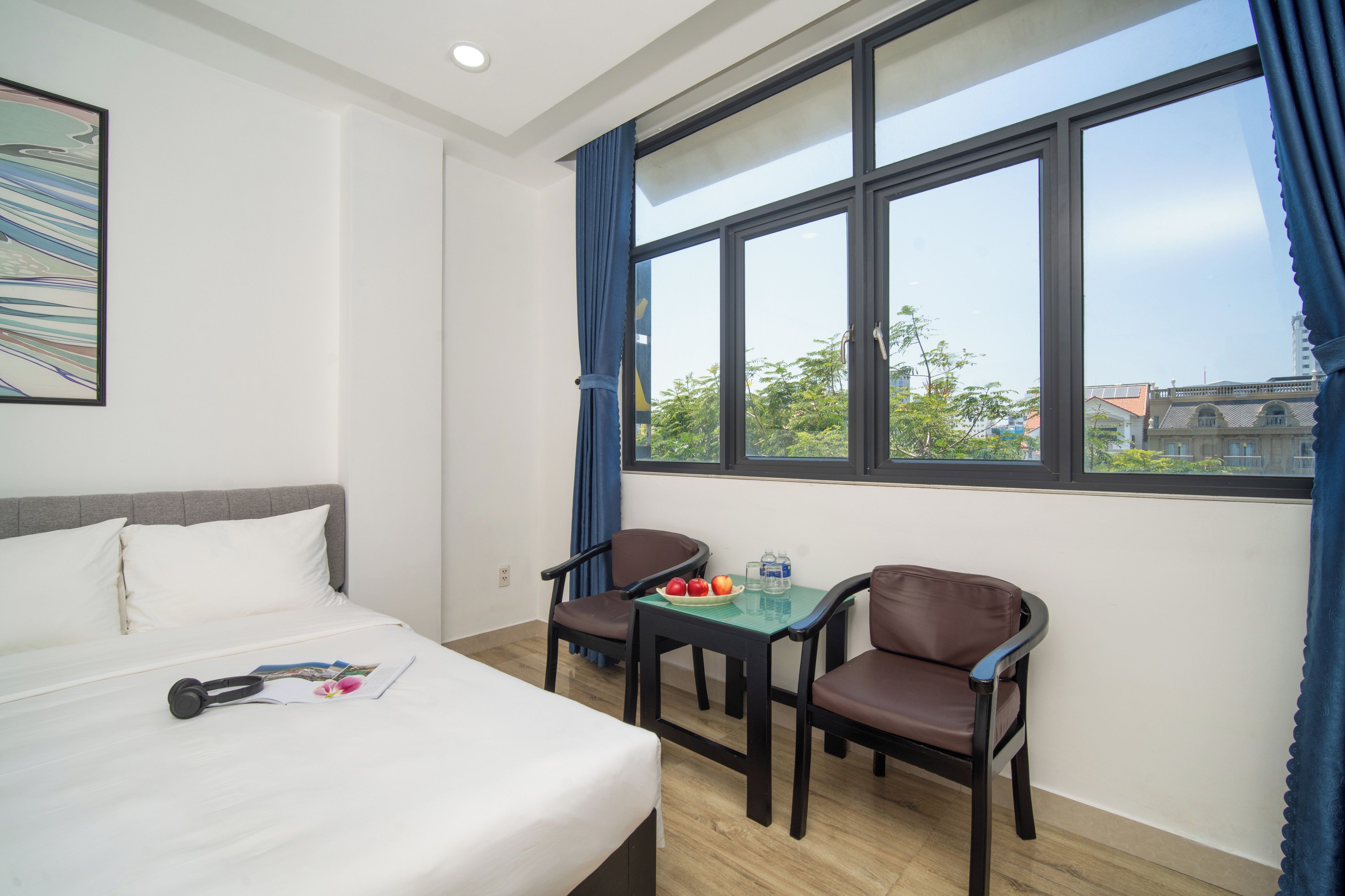 A hotel room with a bed, two chairs, a small table with fruits and water bottles, and a window with curtains.