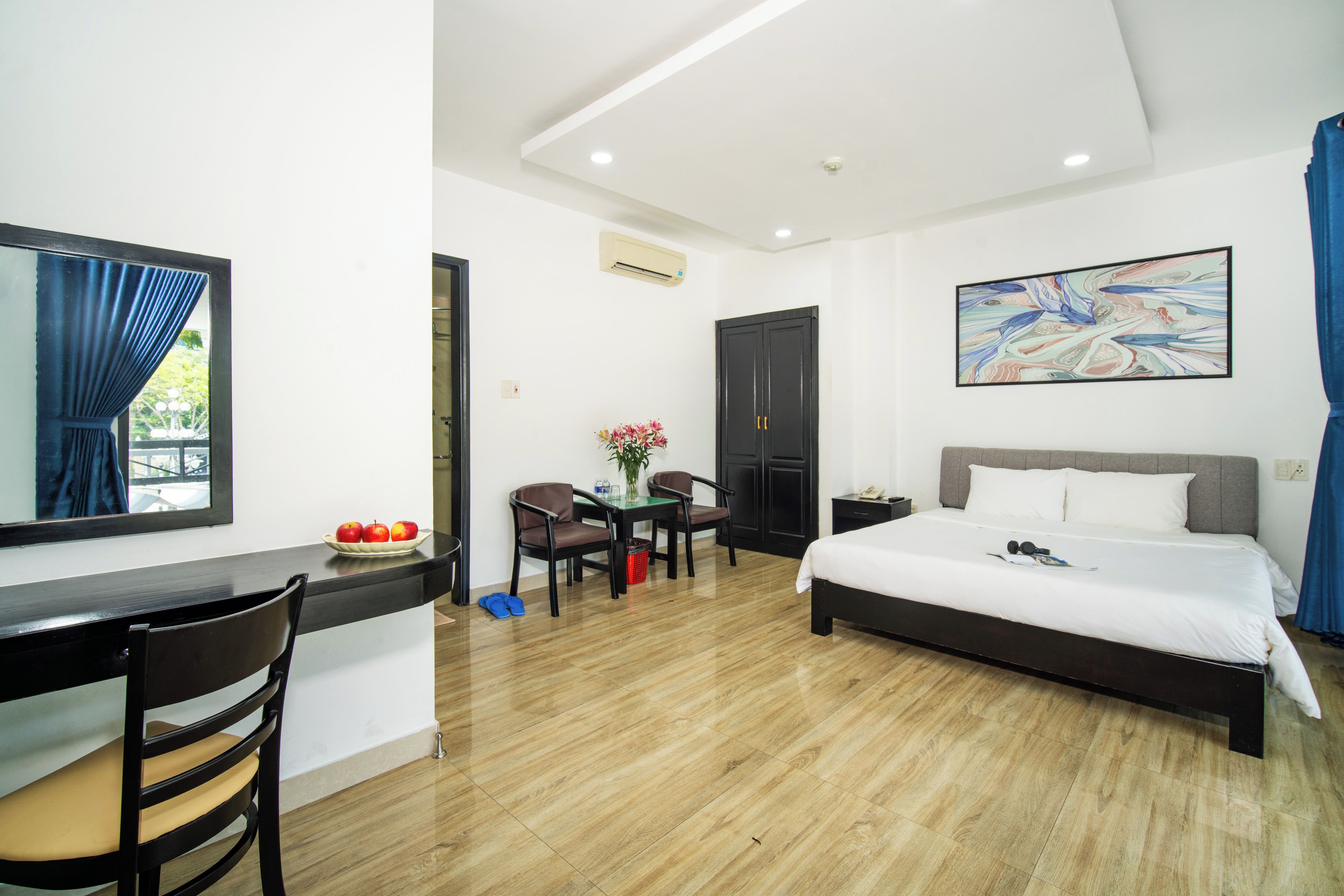 A hotel room with a bed, a desk, a chair, a table with fruit, and a painting on the wall.