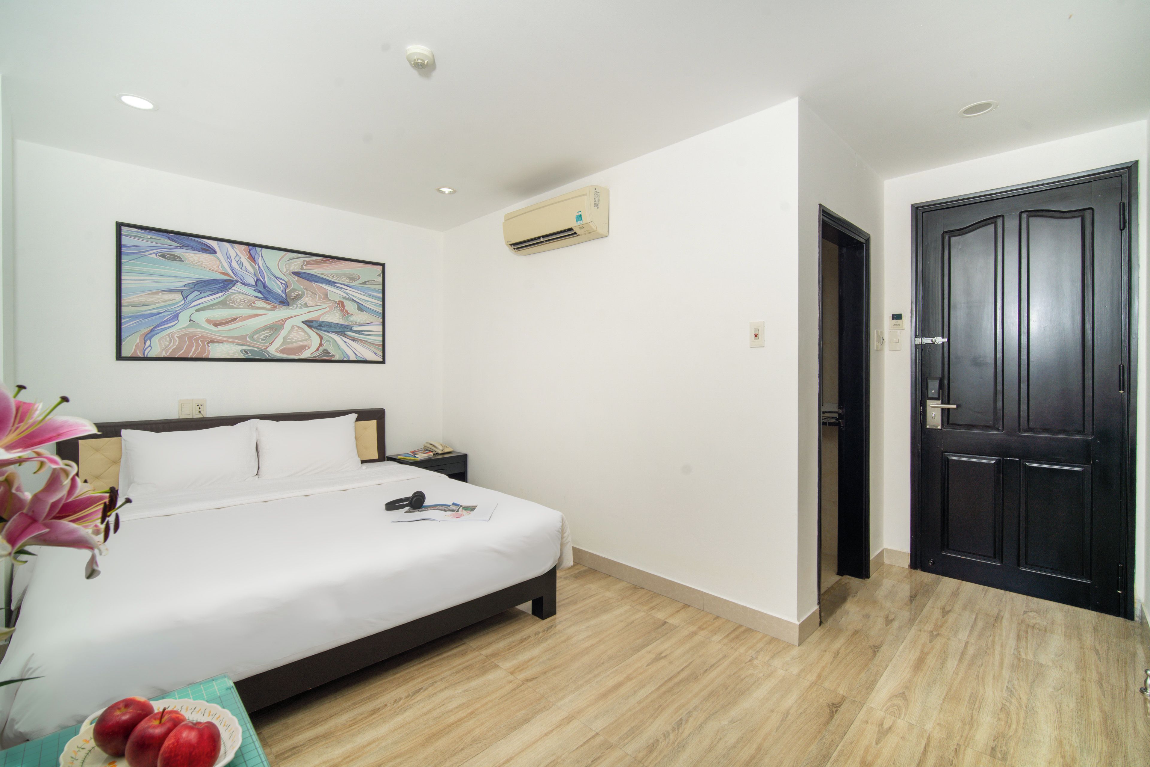 A hotel room with a bed, a painting on the wall, an air conditioning unit, and a door.