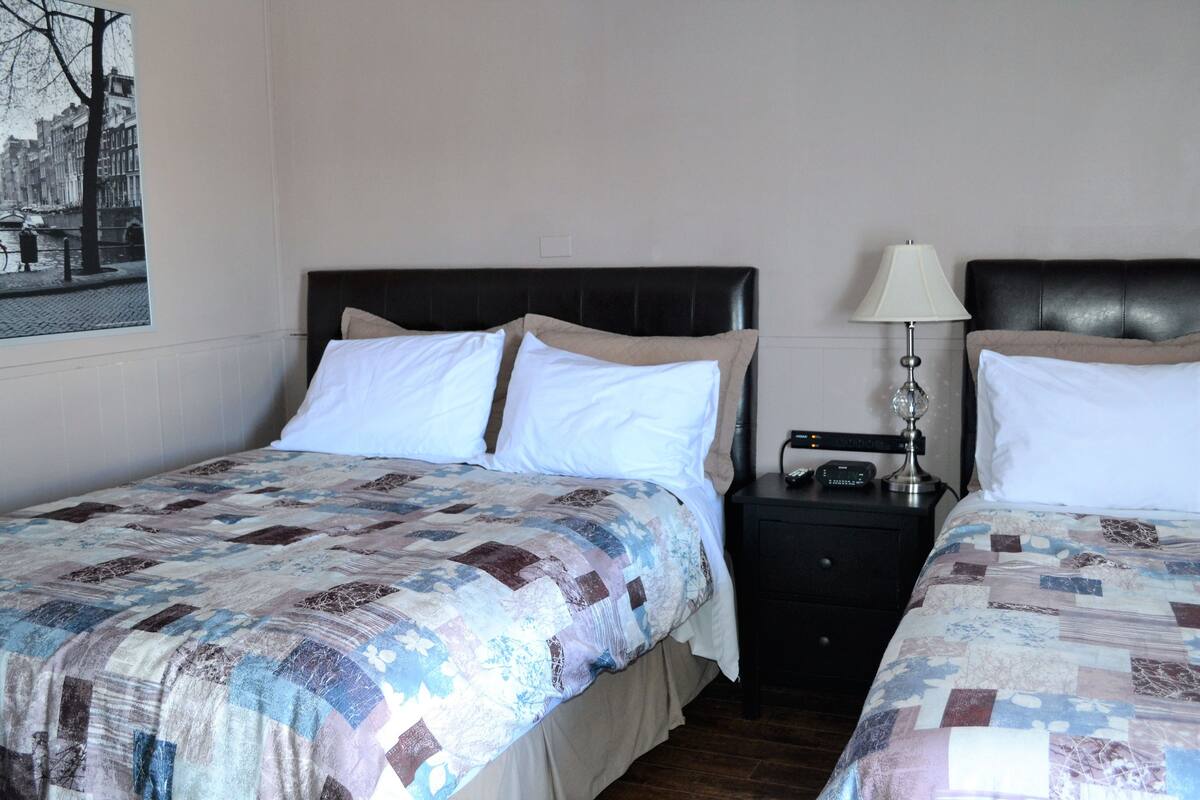 A hotel room with two beds, each with a quilted bedspread and white pillows. There is a nightstand with a lamp and a small table with a clock.