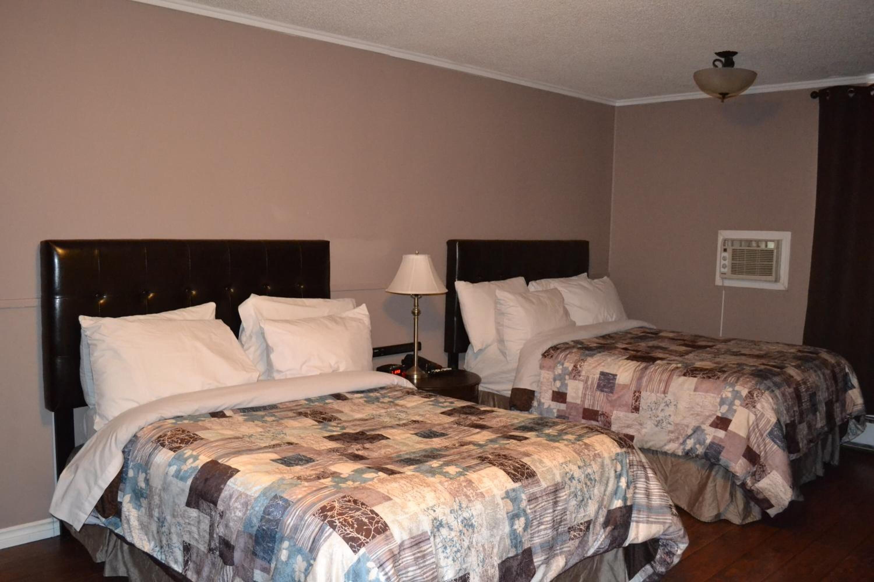 A hotel room with two beds, each with a headboard and a quilted bedspread. There is a nightstand with a lamp between the beds, and a ceiling light fixture above.