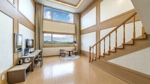 Una habitación amplia con suelo de madera, un escritorio con computadora, una silla, una mesita y vistas al exterior a través de un ventanal grande.
