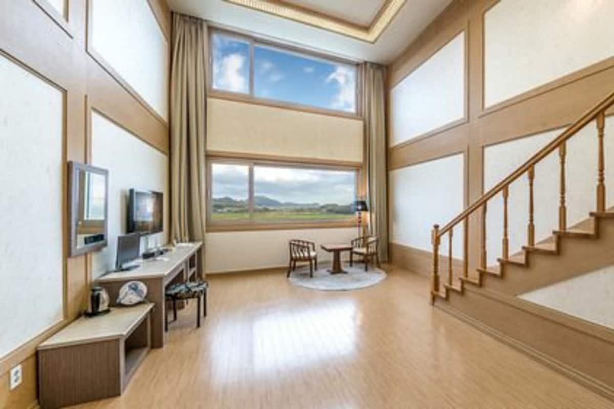 Una habitación amplia con suelo de madera, un escritorio con computadora, una silla, una mesita y vistas al exterior a través de un ventanal grande.
