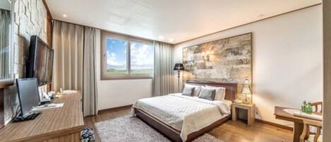 Ein modernes Hotelzimmer mit einem großen Bett, einem Schreibtisch mit Computer, einem Fernseher und Blick ins Freie.