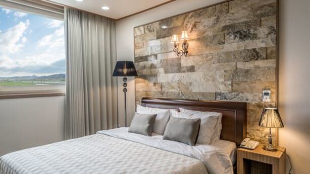 Habitación de hotel con una cama grande, una mesita de noche con lámpara, una lámpara de pared y vistas al exterior a través de una ventana.