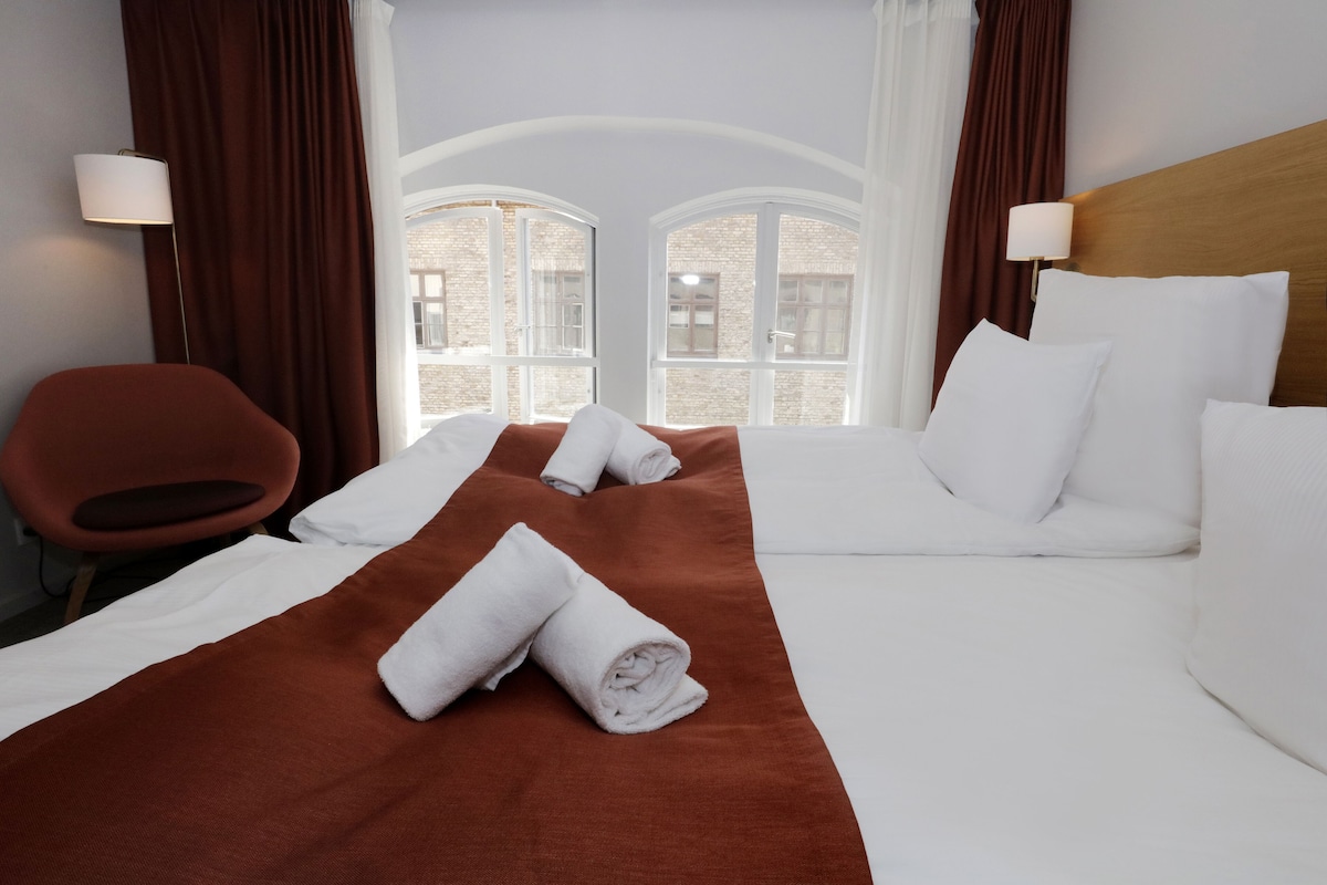 A hotel room with a bed, two pillows, a chair, a window with curtains, and a lamp.