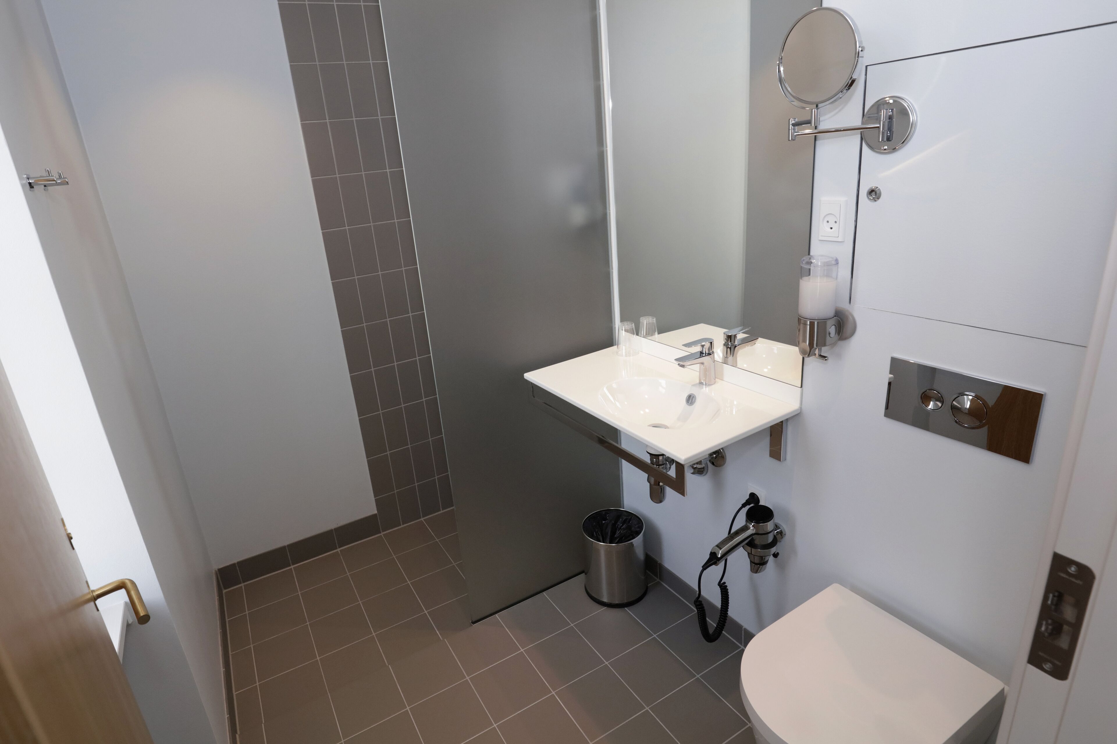 Un bagno moderno con lavandino, specchio e servizi igienici bianchi. Si intravede anche un cestino e uno spazio doccia.