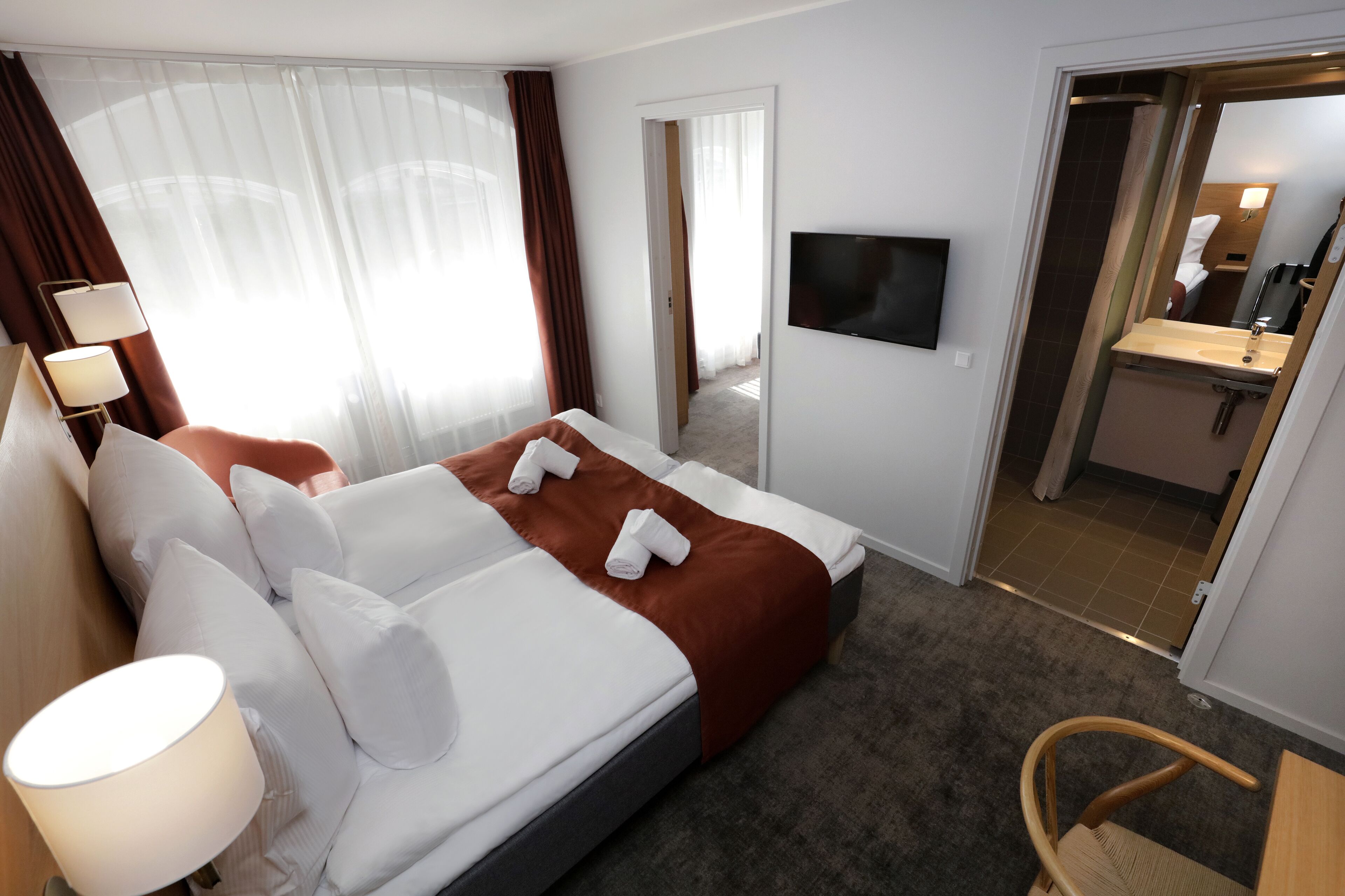 Une chambre d’hôtel avec un lit, un téléviseur fixé au mur, un bureau avec une chaise et une salle de bain visible à travers une porte ouverte.