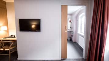 Une chambre d’hôtel dotée d’un téléviseur à écran plat fixé au mur, d’une porte en bois donnant accès à la salle de bain et d’un sol recouvert d’une moquette grise.