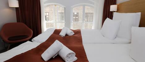Quarto de hotel com uma cama grande, duas luminárias de cabeceira, um cobertor marrom e vista para edifícios através de janelas em arco.