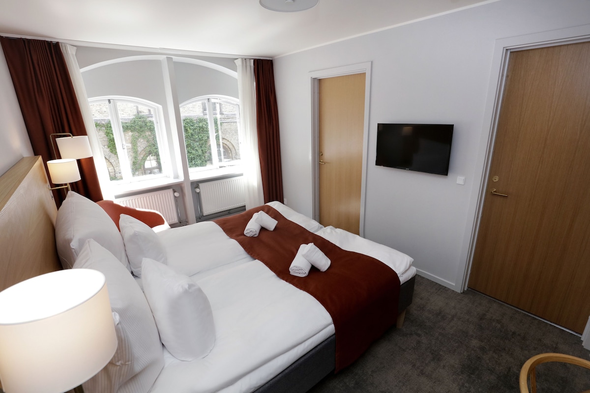 A hotel room with a bed, a TV, a window with curtains, and a door.