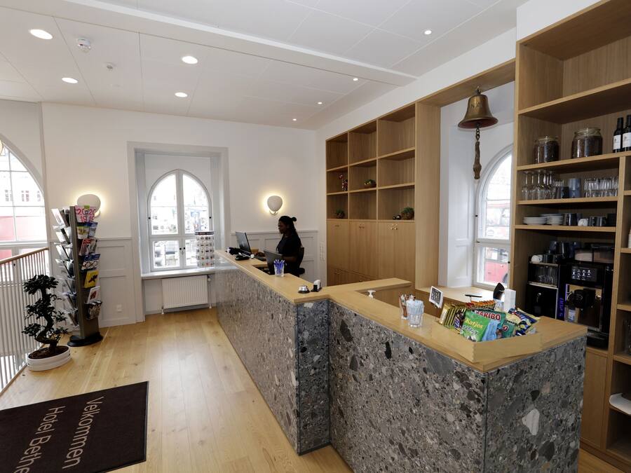 A hotel reception area with a marble counter, wooden shelving with wine bottles, and a view through a window.