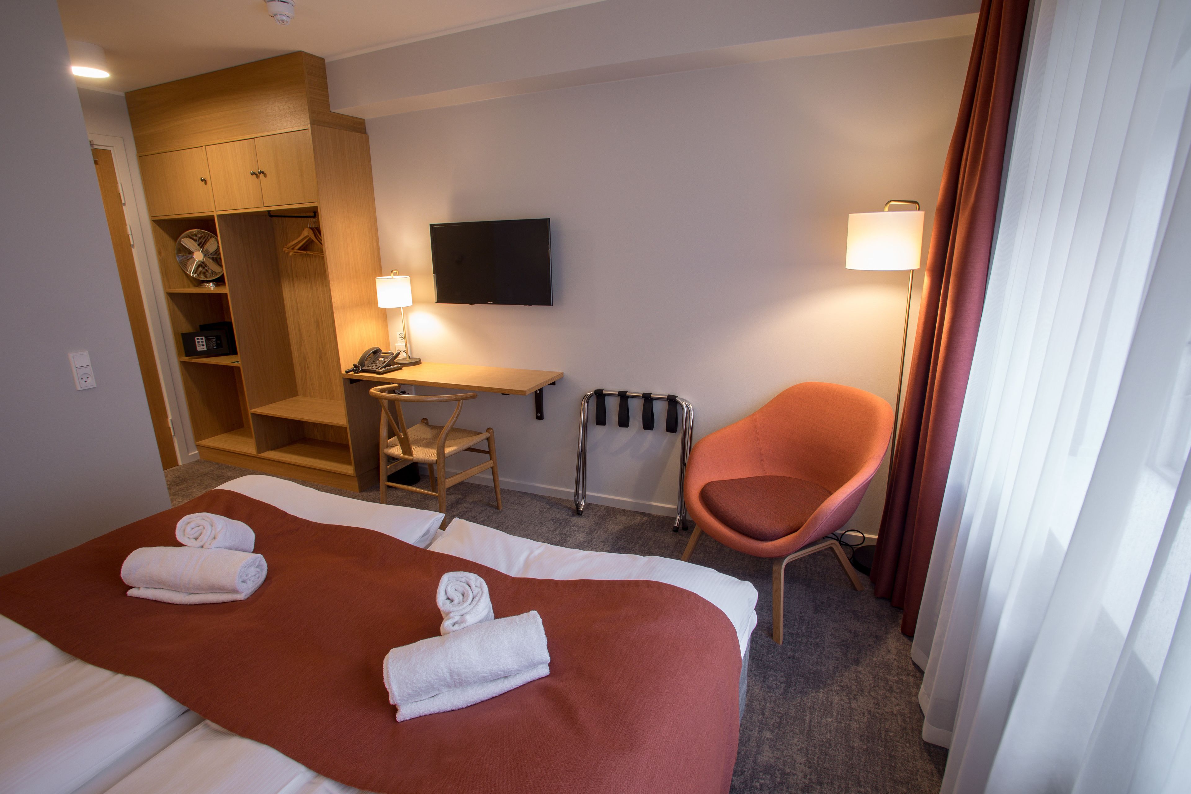 A hotel room with a large bed, a desk, a chair, a TV, and a wardrobe.