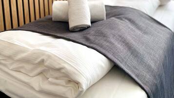 Una cama bien hecha con una manta gris, toallas enrolladas, un cabecero de madera y almohadas blancas.