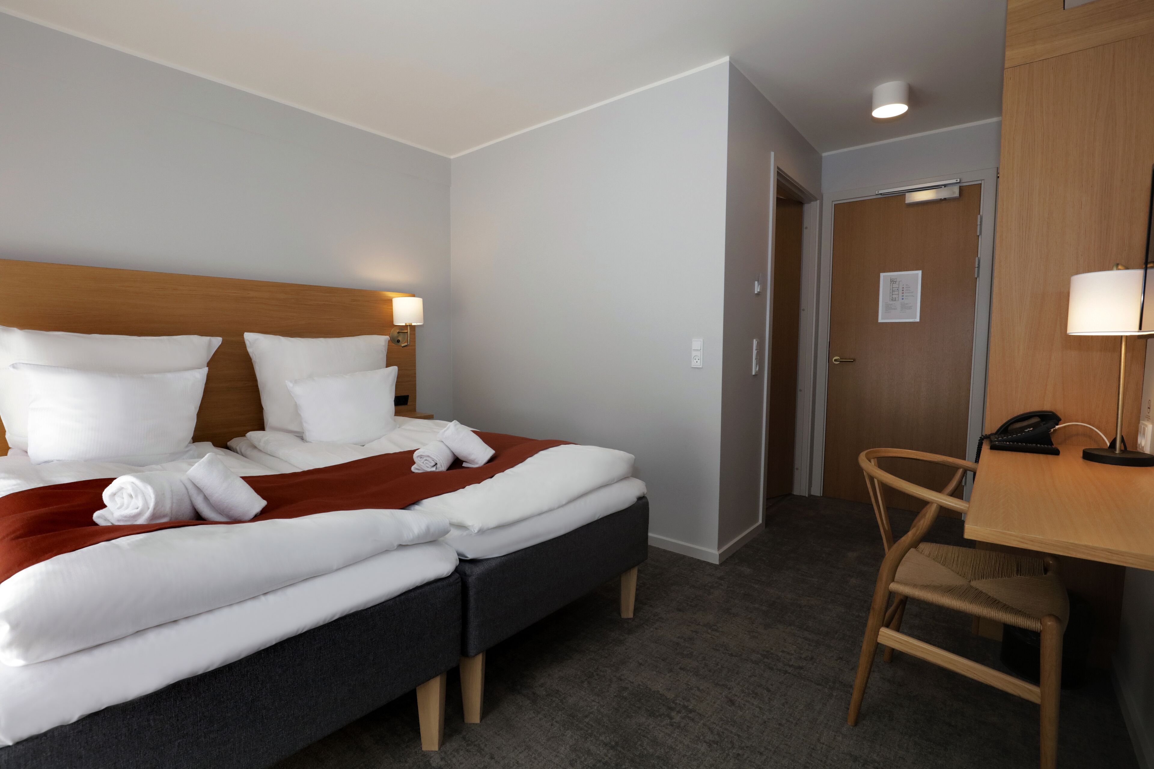 Quarto de hotel com uma cama, duas mesas de cabeceira, uma escrivaninha e uma cadeira, além de uma porta.