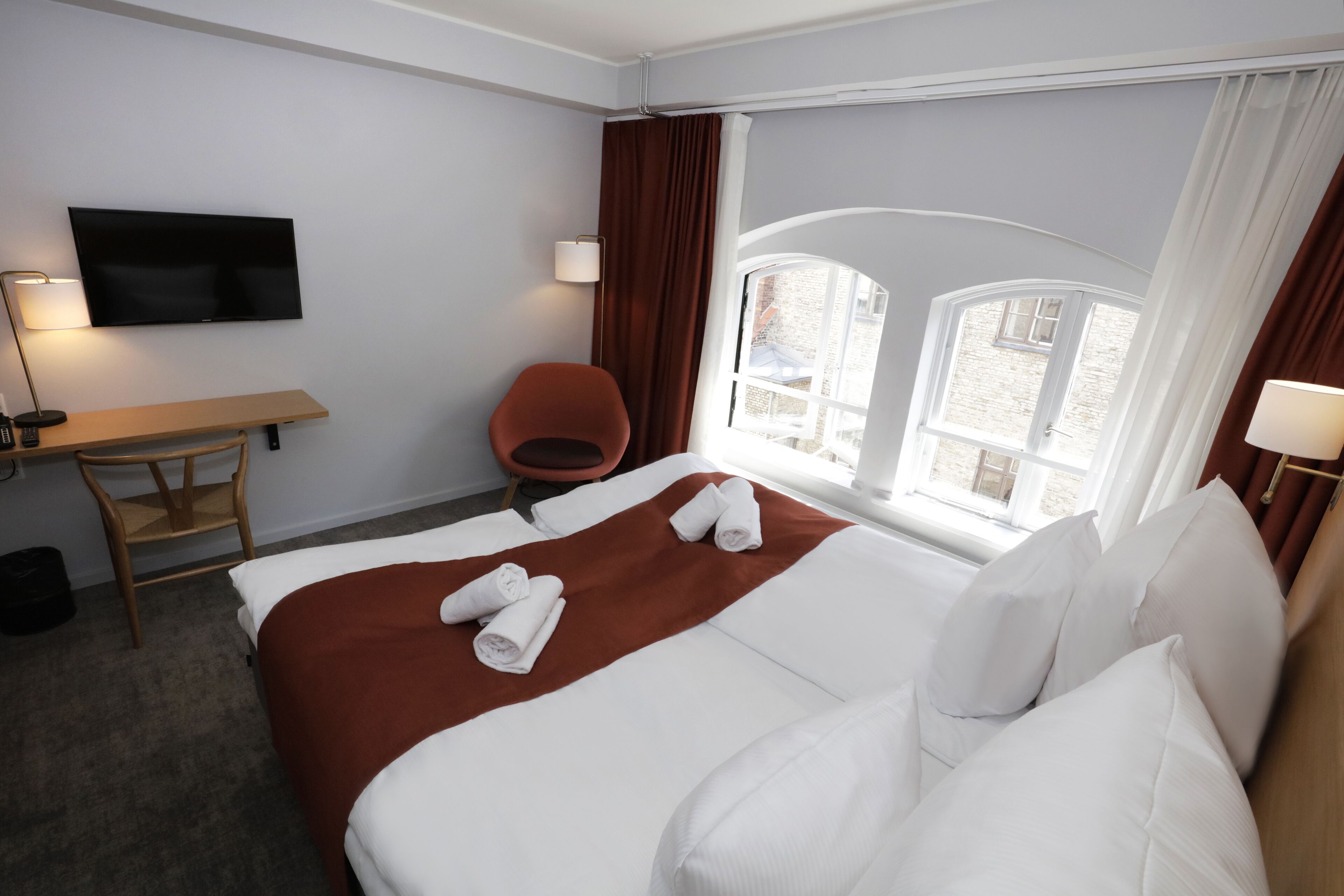 A hotel room with a bed, a desk, a chair, a TV, and a window with curtains.