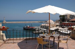 A terrace with a table and chairs, overlooking a harbor with boats and a lighthouse. - Belmondo Hotel (Chania)