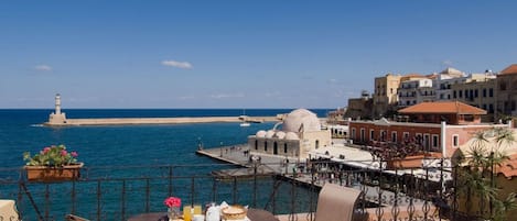 A terrace with a table set for breakfast overlooking a coastal town, a lighthouse, and a historic building with domes.