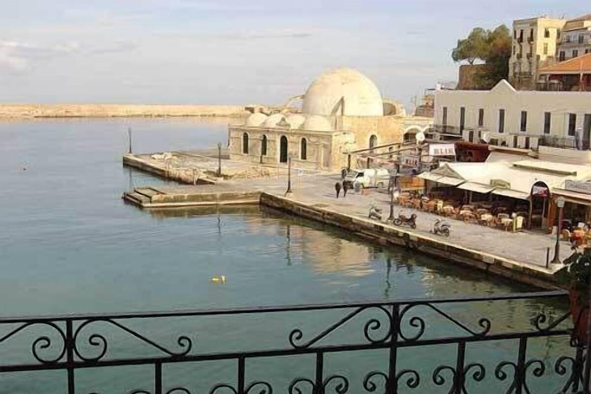 Vista costera con un edificio blanco con cúpula, un muelle y un área de bar con mesas al aire libre.