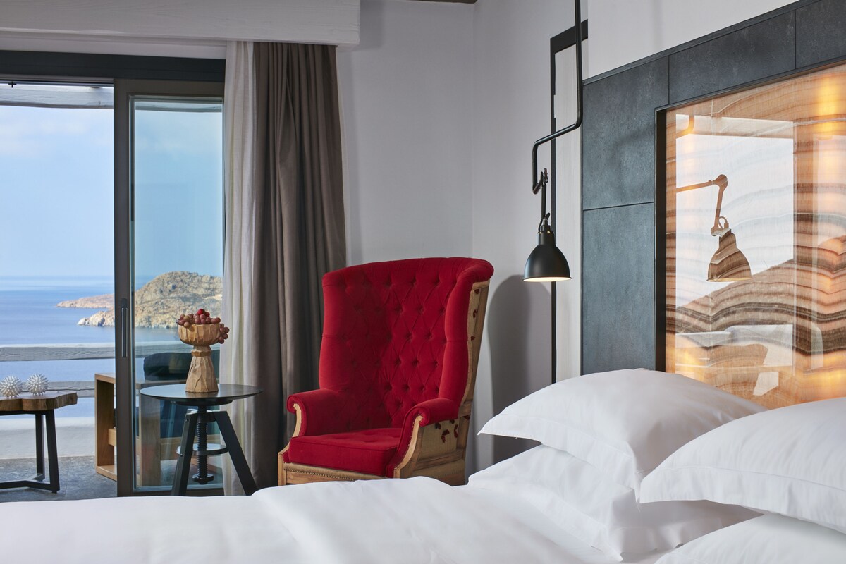 A hotel room with a bed, a red armchair, a small table with a fruit bowl, and a large window offering a view of the sea.