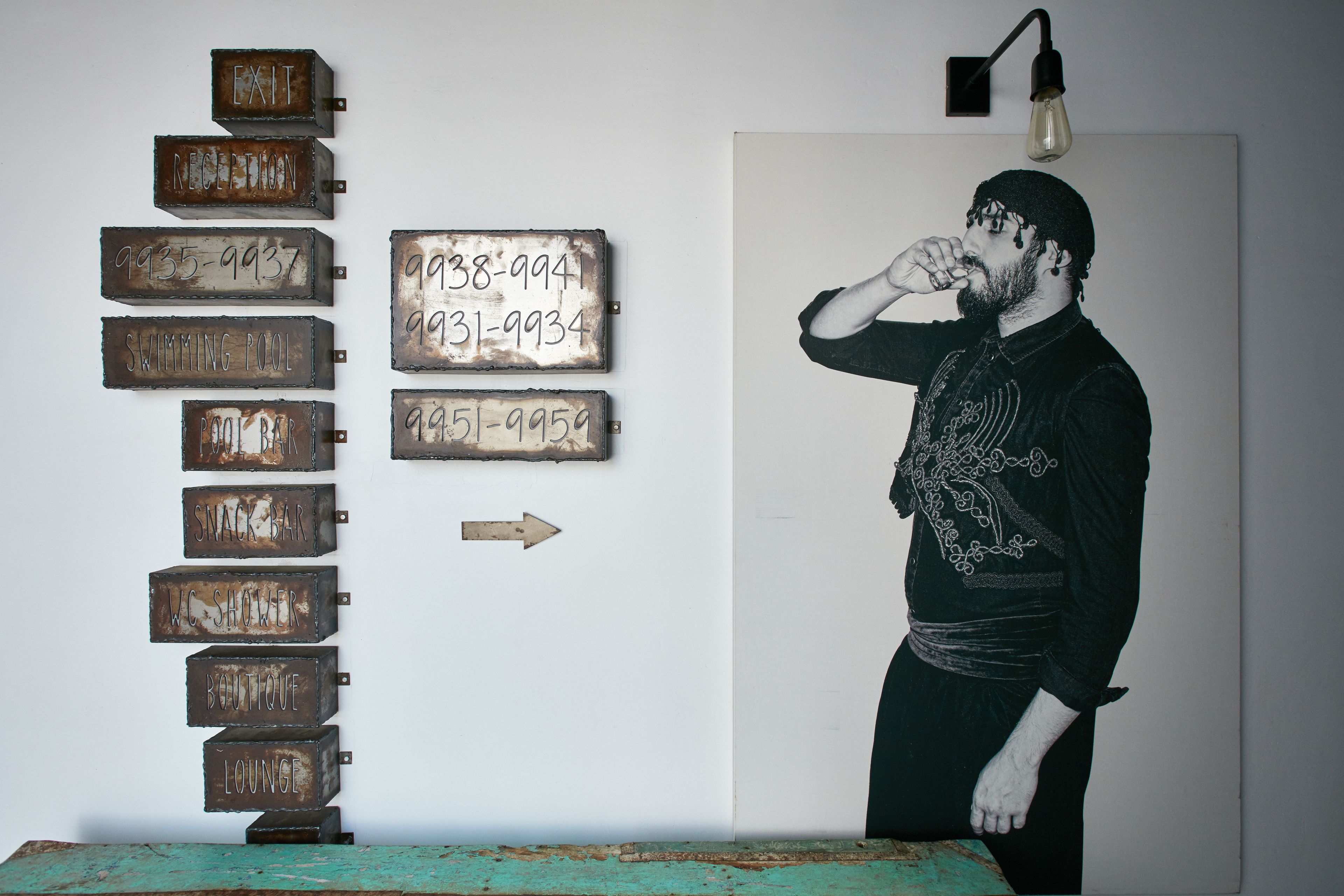 A wall with metal signs indicating various locations such as 'EXIT', 'RECEPTION', 'SWIMMING POOL', 'SNACK BAR', 'WC SHOWER', 'BOUTIQUE', and 'LOUNGE'.