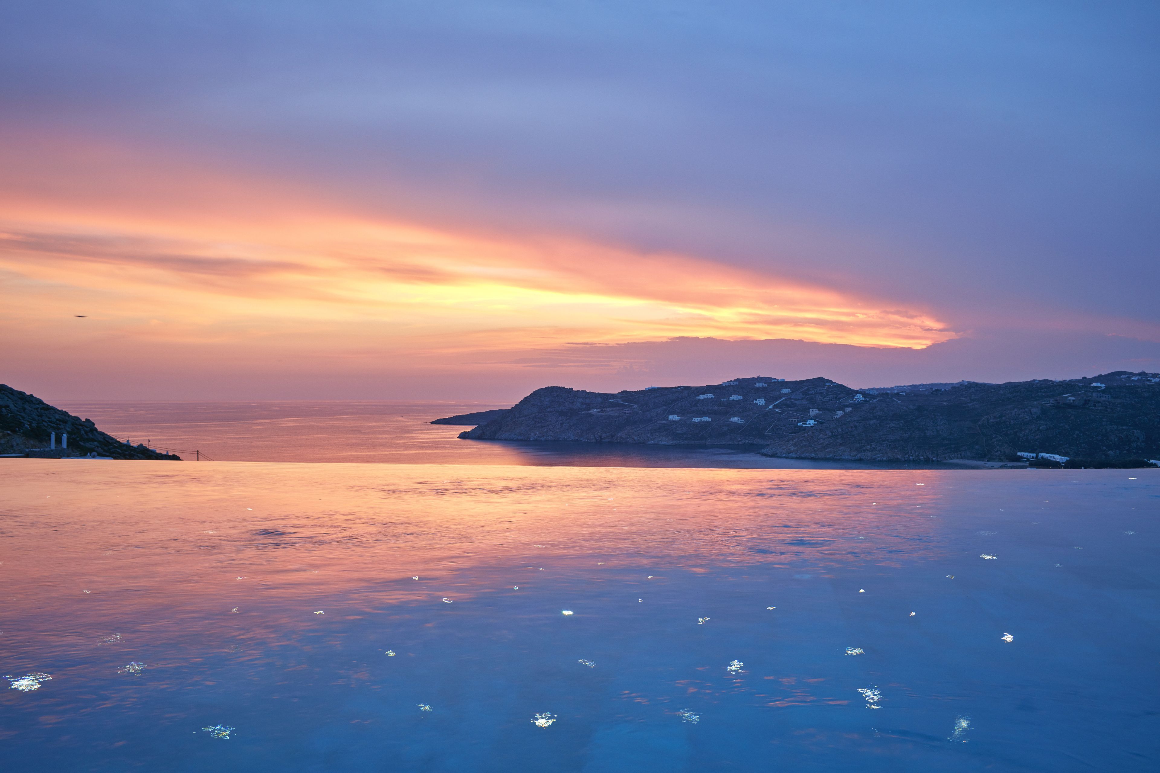 Eine Küstenlandschaft mit Sonnenuntergang über dem Wasser, felsigem Gelände und einer entfernten Ansicht einer kleinen Siedlung.
