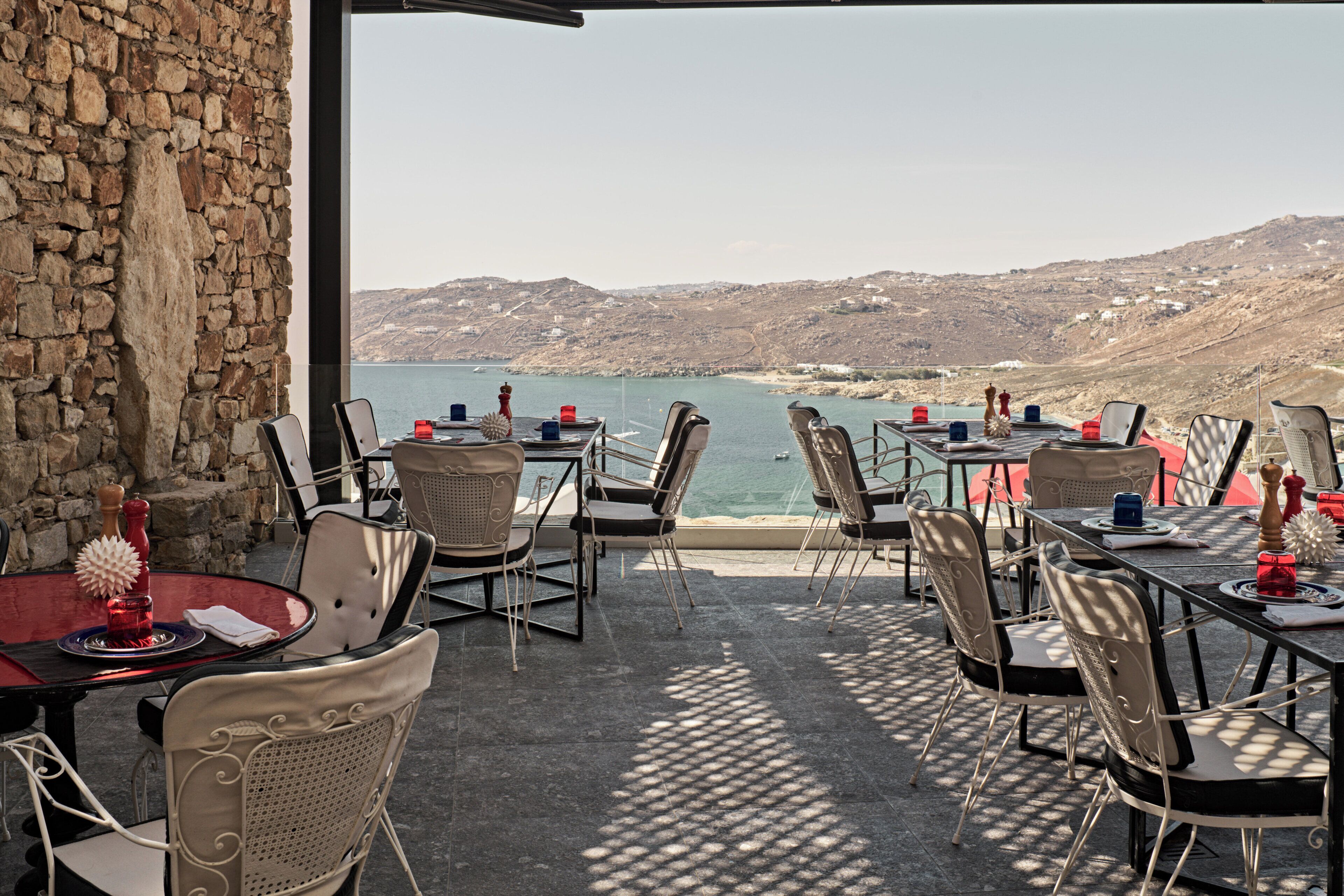 An outdoor dining area with stone walls, tables, and chairs, overlooking a coastal landscape.