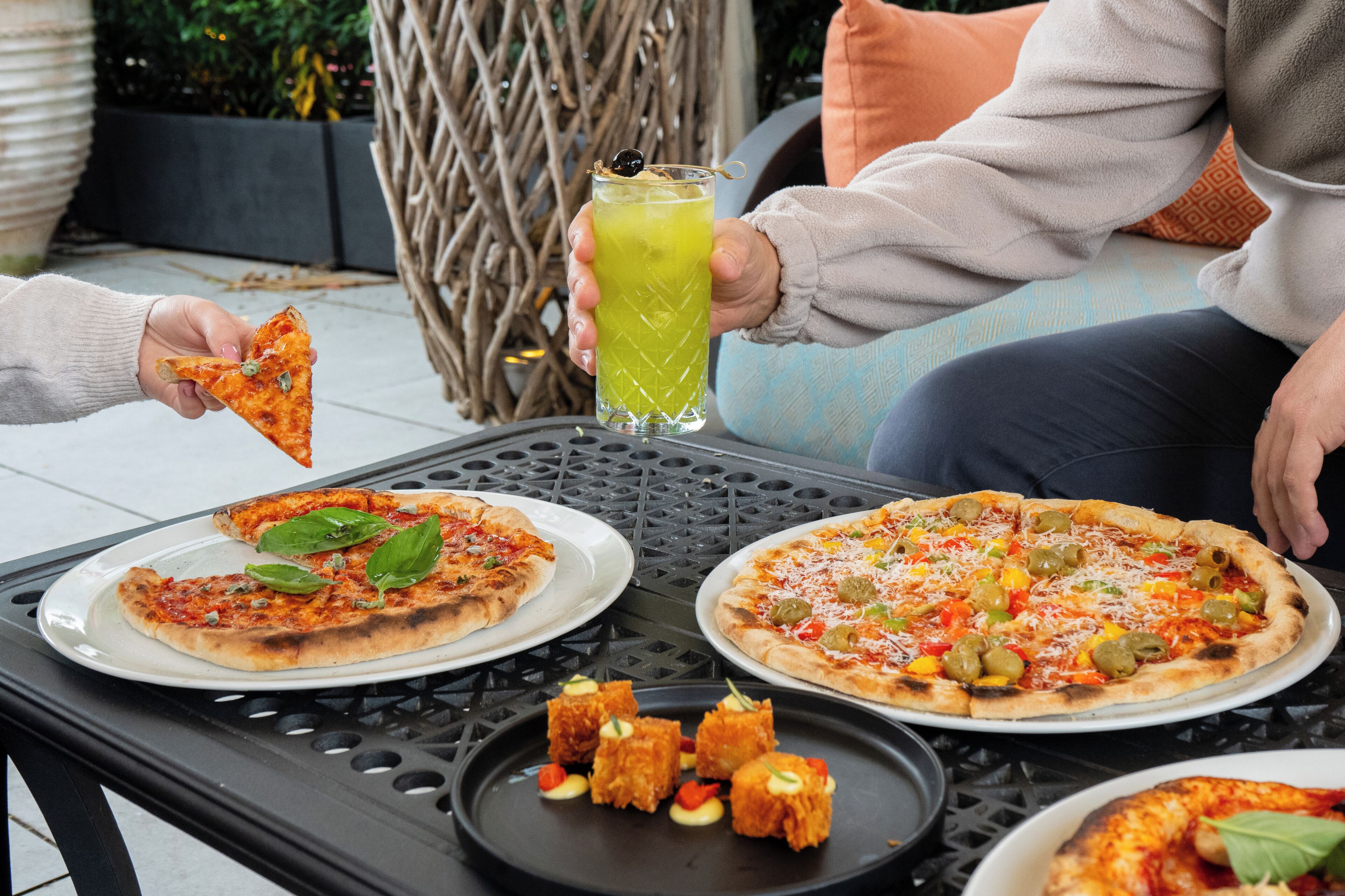 A dining table with two pizzas, a glass of beverage, and a plate with appetizers.