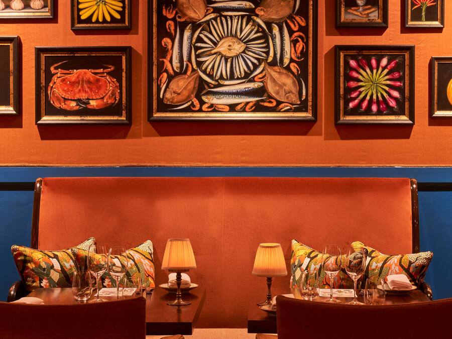 A dining area with two tables, each with a lamp and a glass of water. The walls are adorned with framed artworks depicting various subjects.