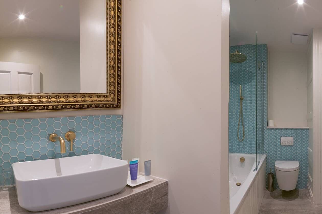 Une salle de bain avec un lavabo blanc, un miroir à cadre doré et une zone de douche carrelée de carreaux bleus.