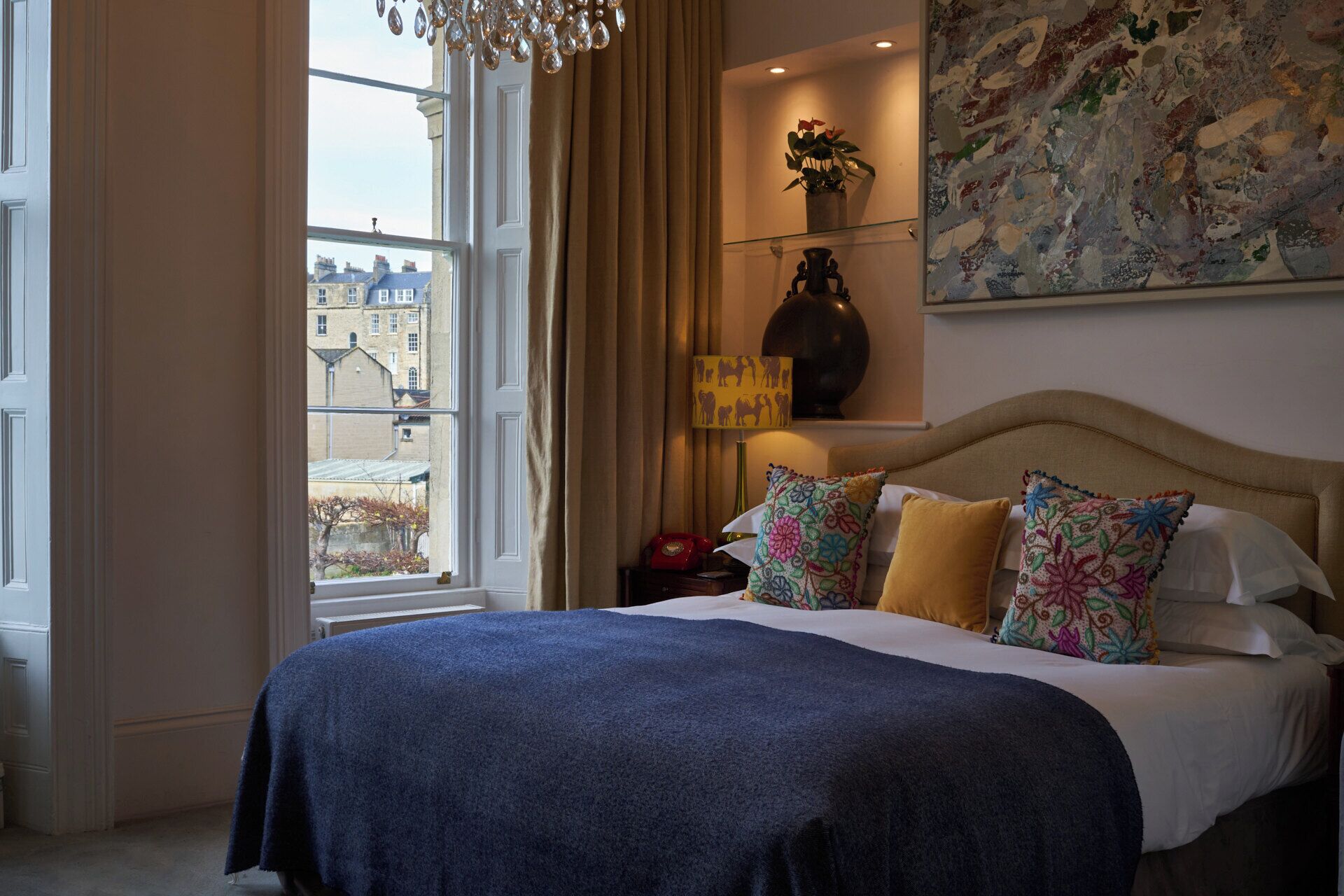 A hotel room with a large bed, a nightstand, a lamp, a vase, and a painting on the wall.