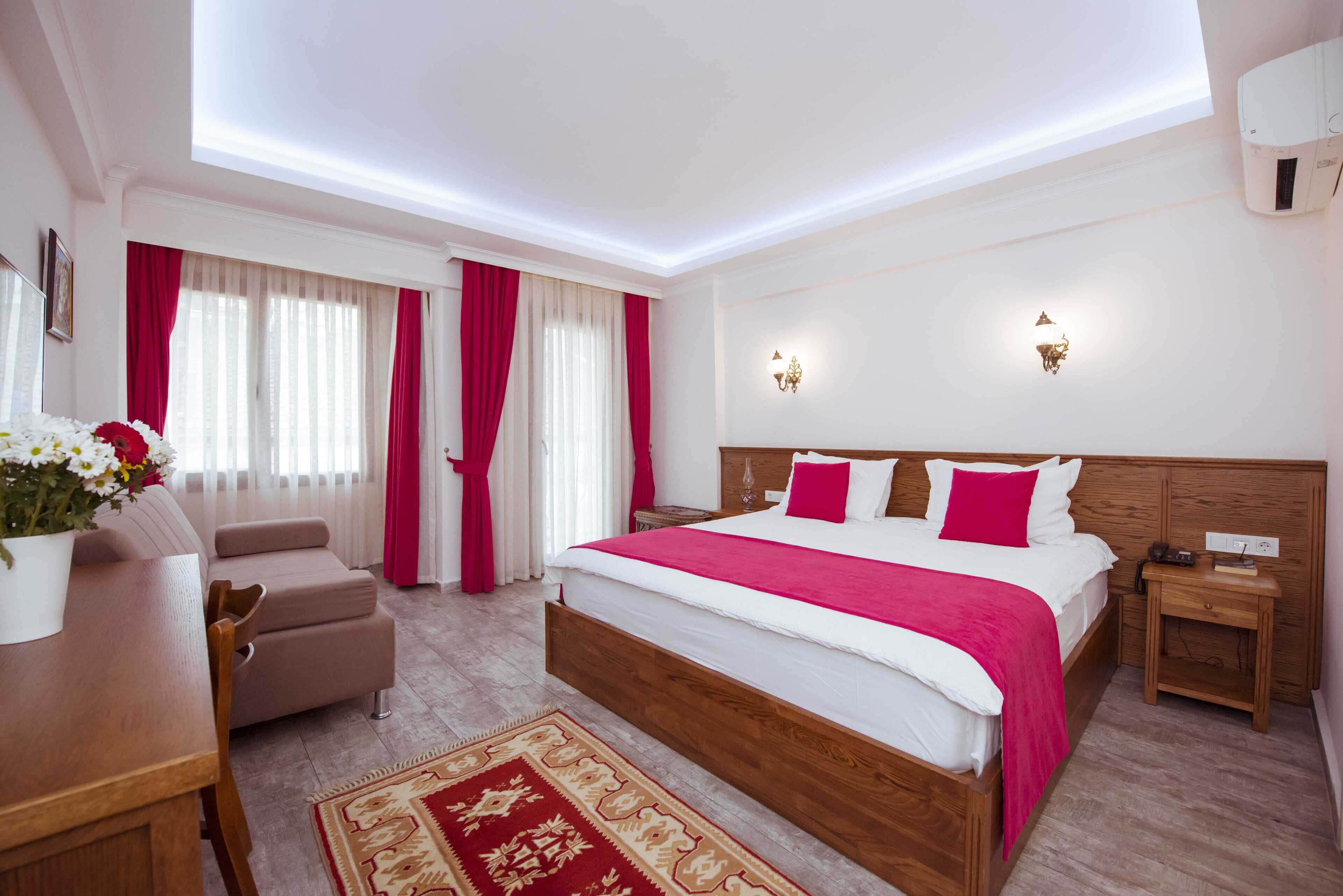 A hotel room with a large bed, a wooden headboard, a bedside table, a nightstand, a desk, a chair, a red and beige patterned rug, and a window with red curtains.