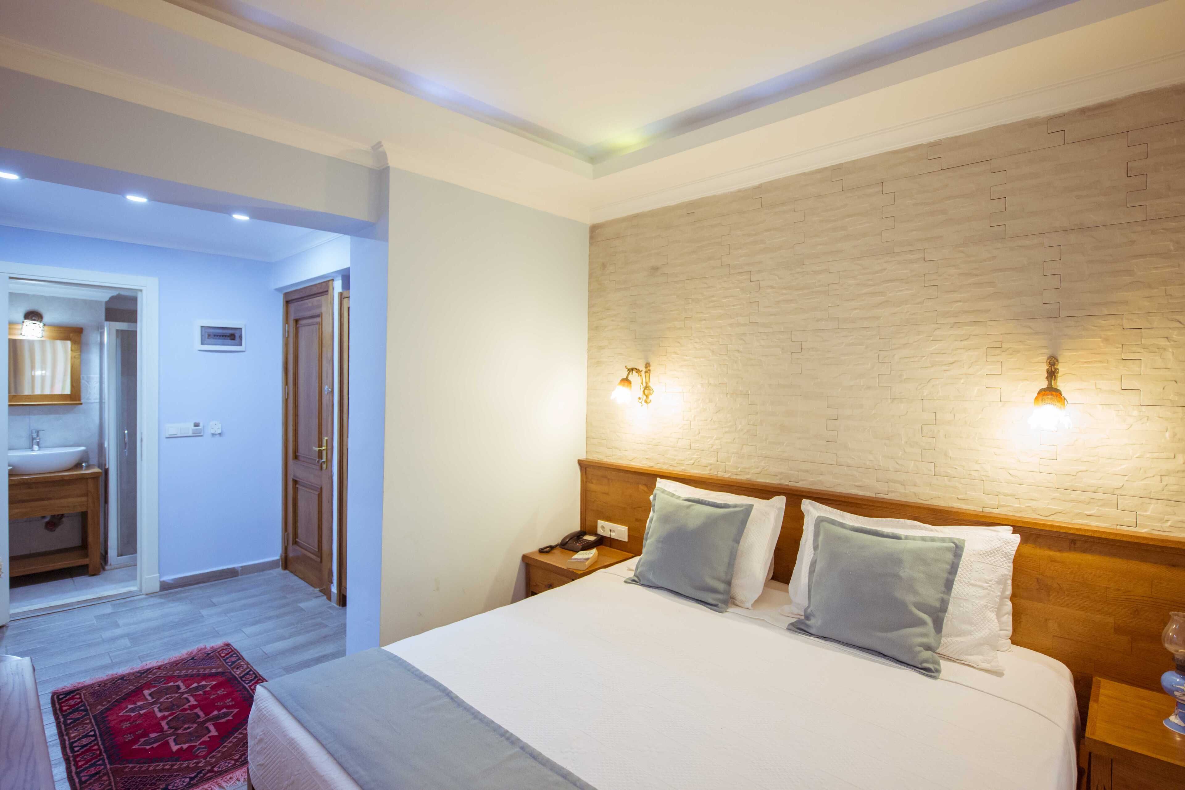 A hotel room with a large bed, wooden headboard, two bedside tables, a nightstand, a red rug, a bathroom with a sink and mirror, and a brick-patterned wall.