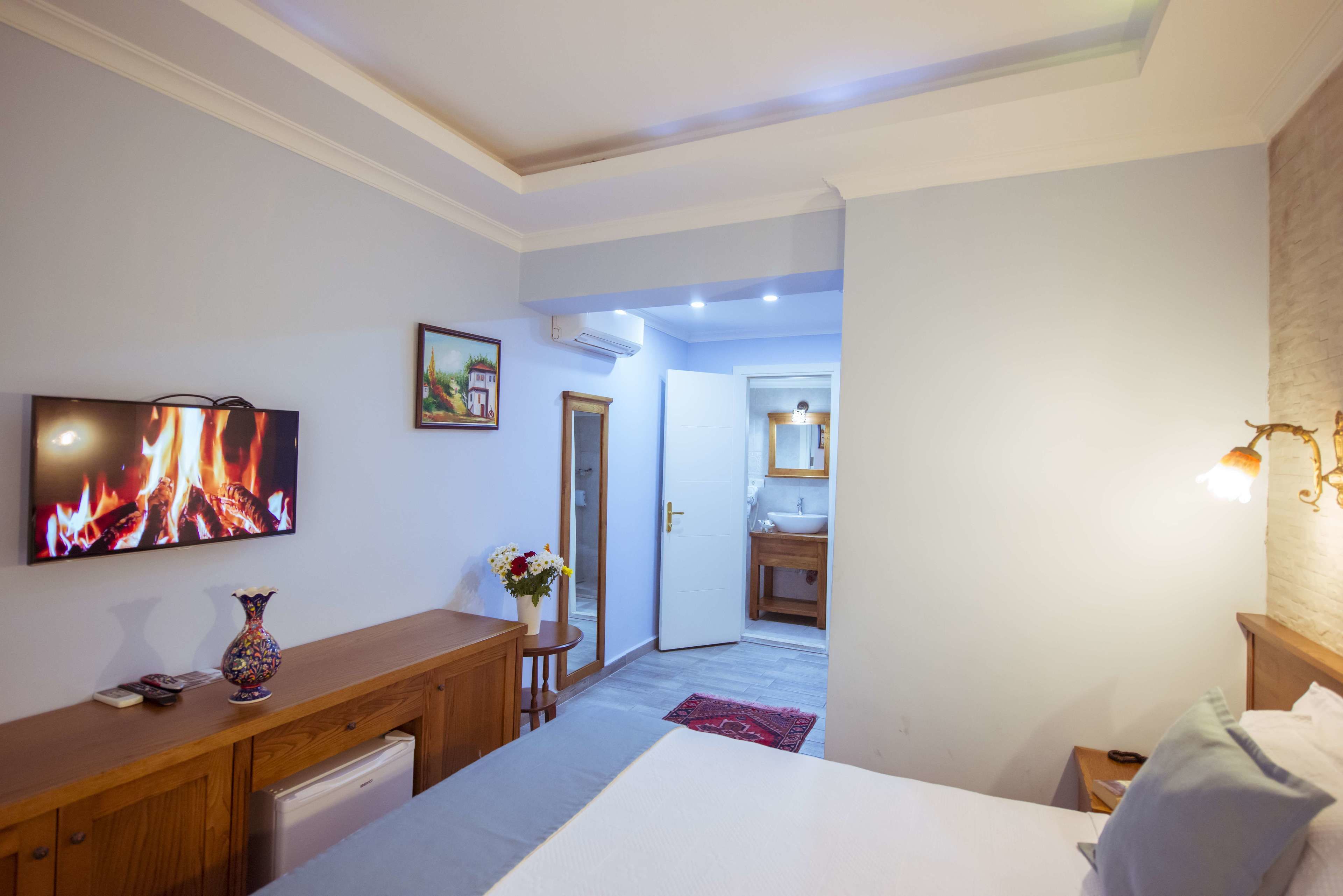 Habitación de hotel con cama, escritorio, televisor mostrando un fuego, baño visible a través de una puerta abierta y una pequeña mesa con un jarrón de flores.