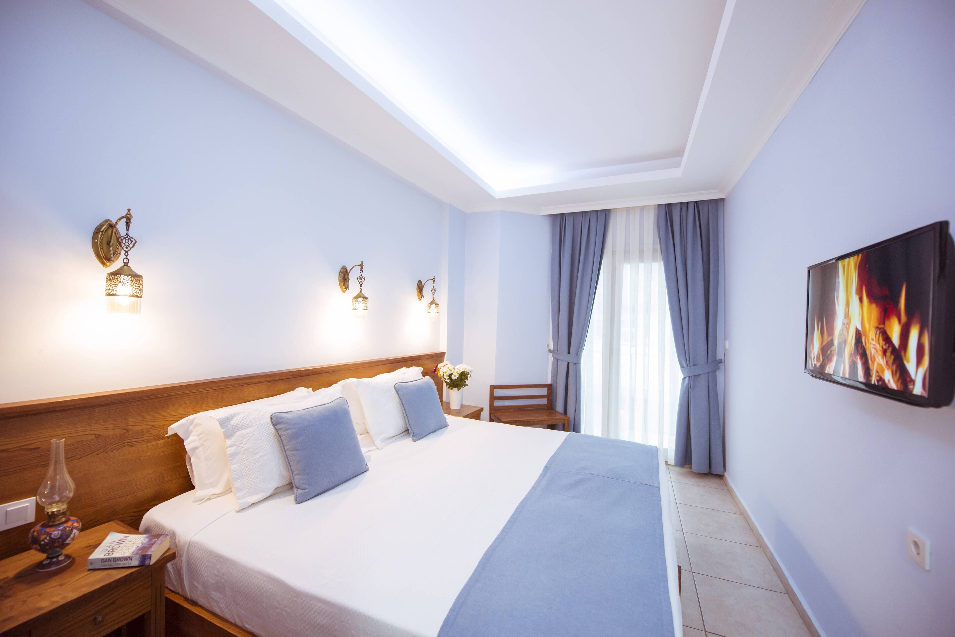A hotel room with a large bed, a nightstand with a lamp, a wall-mounted TV, and a window with curtains.
