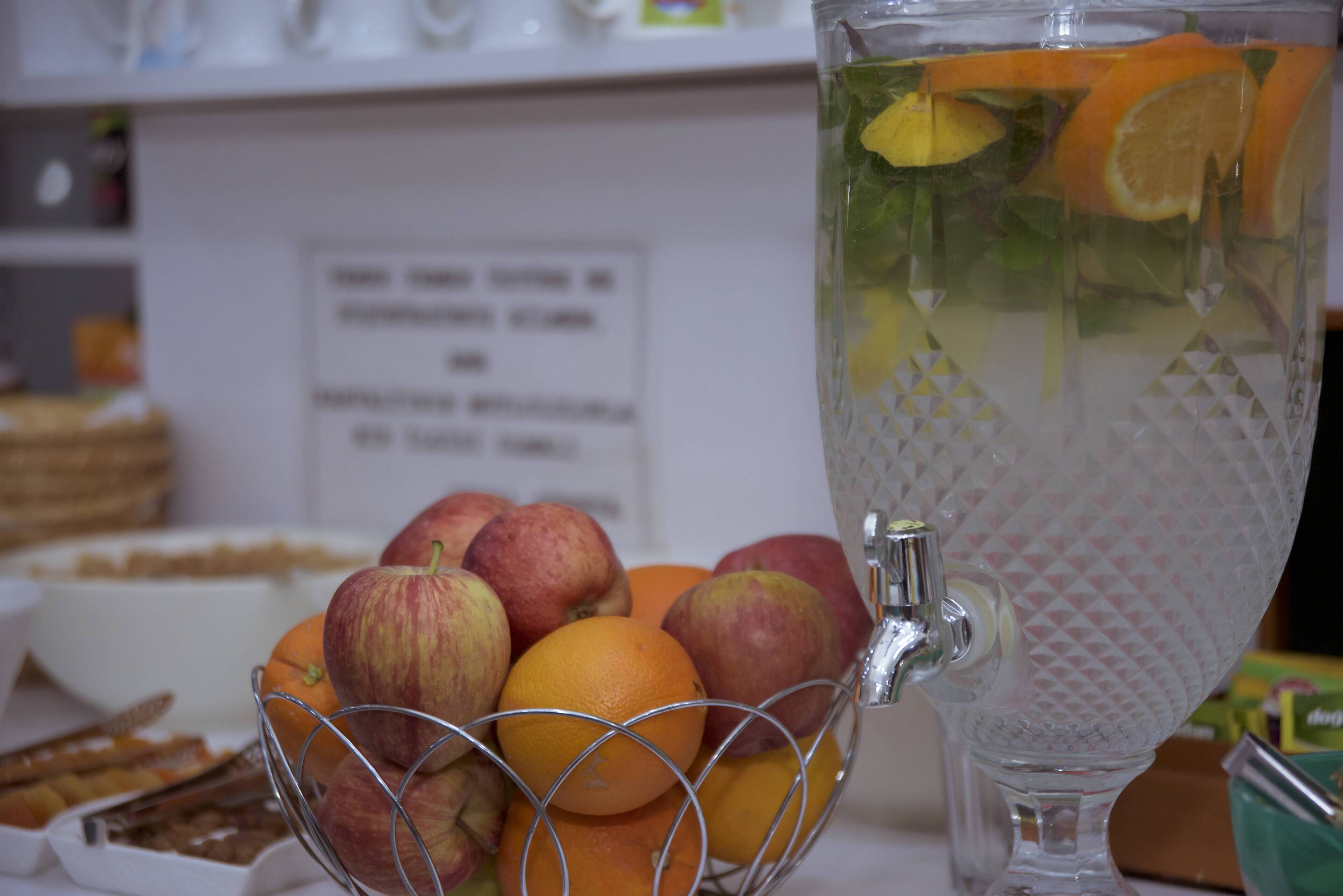 Un frutero con manzanas y naranjas, una jarra de vidrio con rodajas de fruta, y un cartel al fondo.